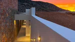 A modern staircase with a stone wall on one side and a white wall on the other, leading to a terrace with a sunset view. - Maison Meltem (Malevizi)