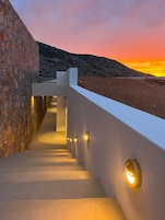 Una escalera moderna con un muro de piedra a un lado y un muro blanco al otro, que lleva a una terraza con vistas al atardecer.