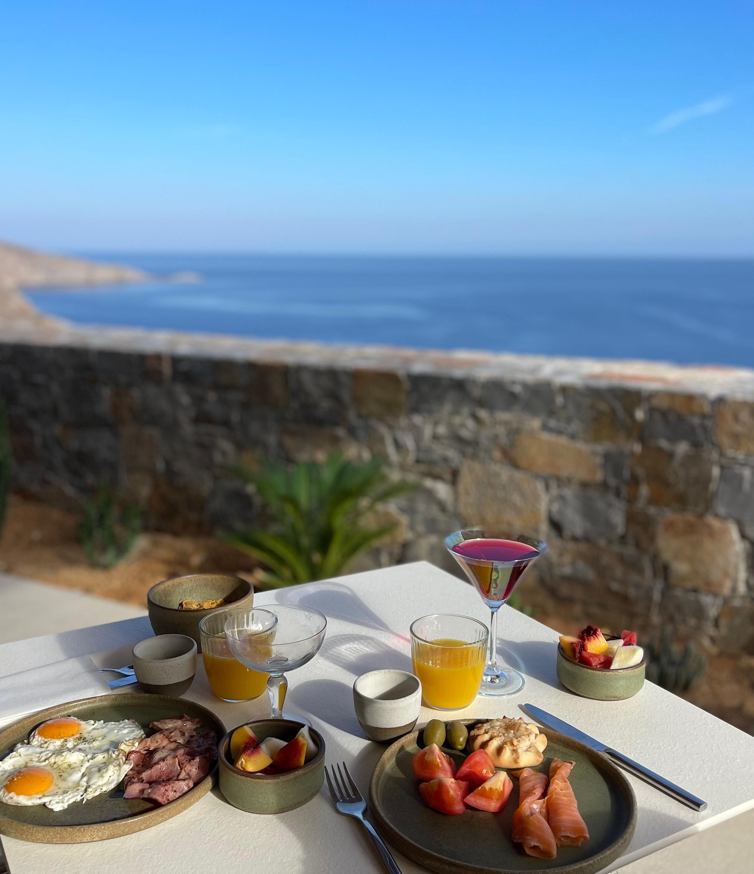 A breakfast setup with eggs, meats, fruits, and drinks on a table overlooking the sea.