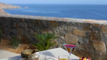 A breakfast setup with eggs, meats, fruits, and drinks on a table overlooking the sea.