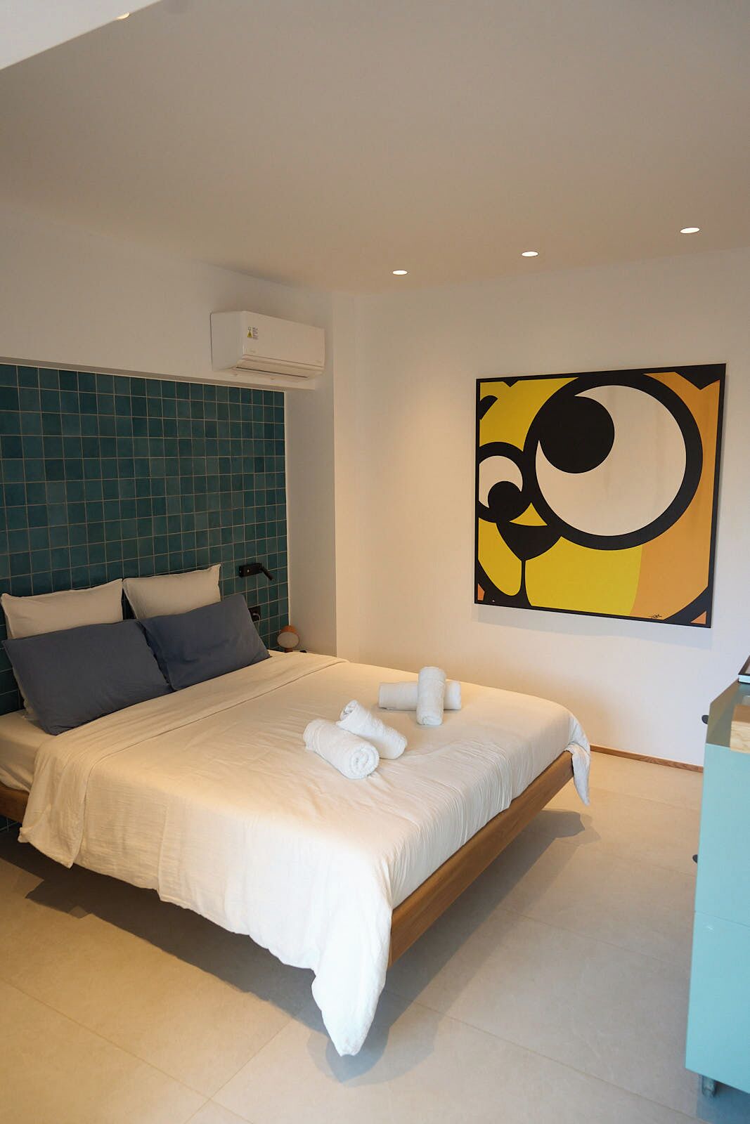 A modern hotel room with a large bed, a teal tiled headboard, a wall-mounted air conditioner, and a large abstract painting.