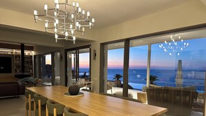 A modern dining area with a wooden table and green chairs, a chandelier, and a view of the ocean through large windows. - Maison Meltem (Malevizi)