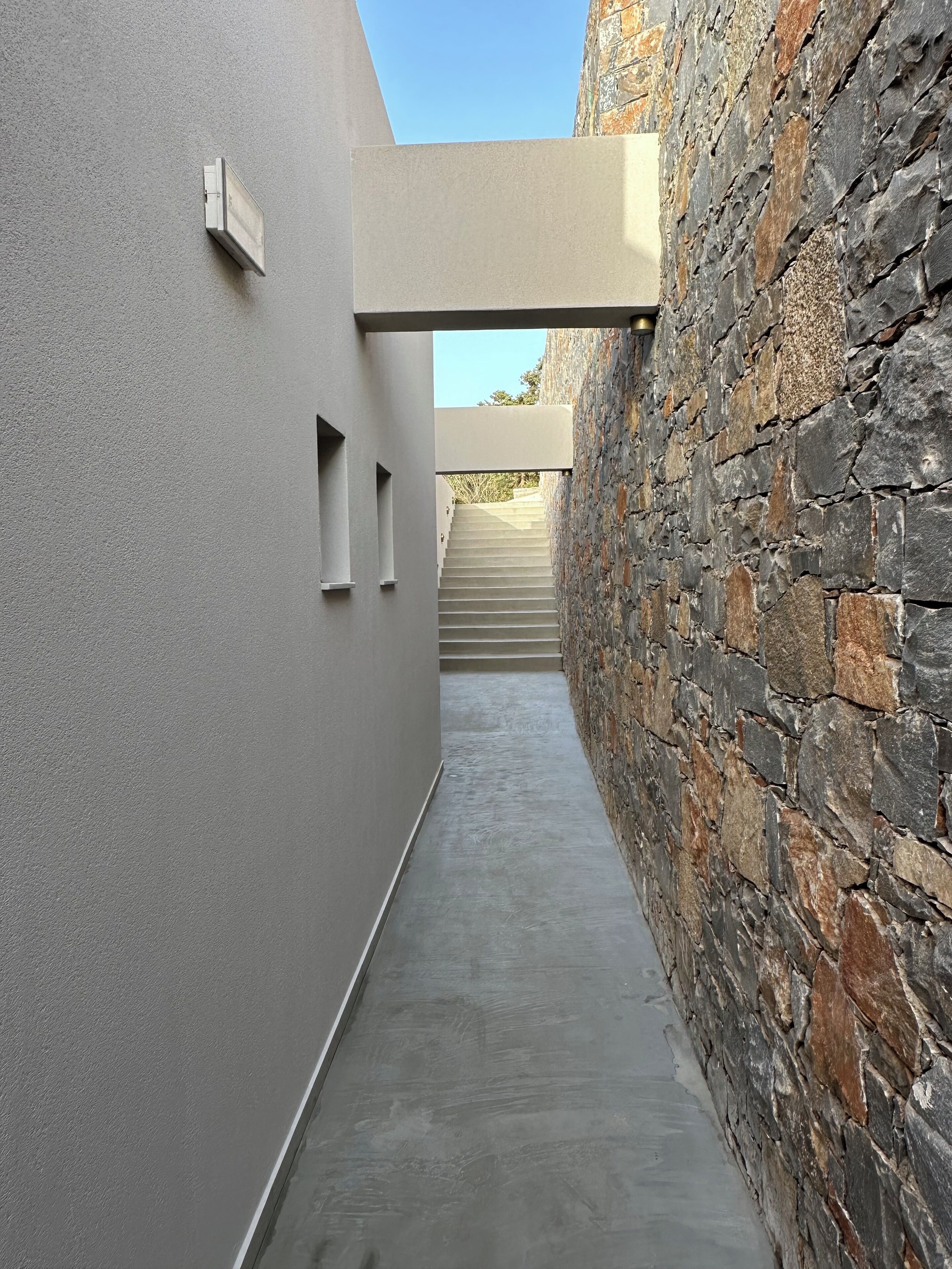 Un couloir étroit, doté d’un sol en béton, d’un mur en pierre d’un côté et d’un mur lisse de l’autre. Un escalier mène à un niveau supérieur.