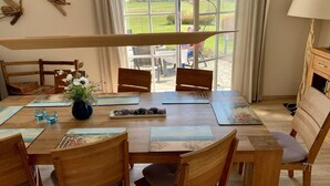 Ein Essbereich mit einem Tisch für vier Personen, Holzstühlen, einer Vase mit Blumen und Blick auf eine grüne Landschaft durch große Fenster.