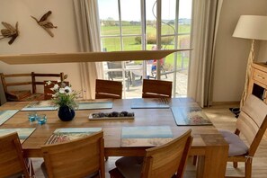 Ein Essbereich mit einem Tisch für vier Personen, Holzstühlen, einer Vase mit Blumen und Blick auf eine grüne Landschaft durch große Fenster.