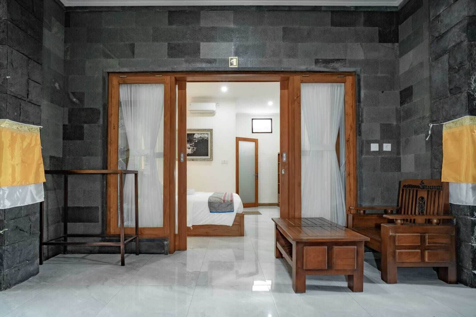 A room with a bed, a wooden bench, a chair, and a small table. There is a door leading to another room.