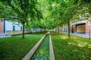 A tree-lined pathway with a narrow water feature, flanked by buildings with brick and white facades.