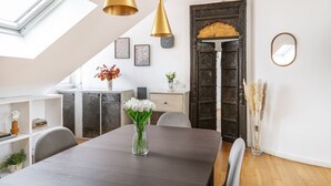 Een eetruimte met een donker houten tafel, witte stoelen en een vaas met witte bloemen. Boven de tafel hangen twee gouden hanglampen.