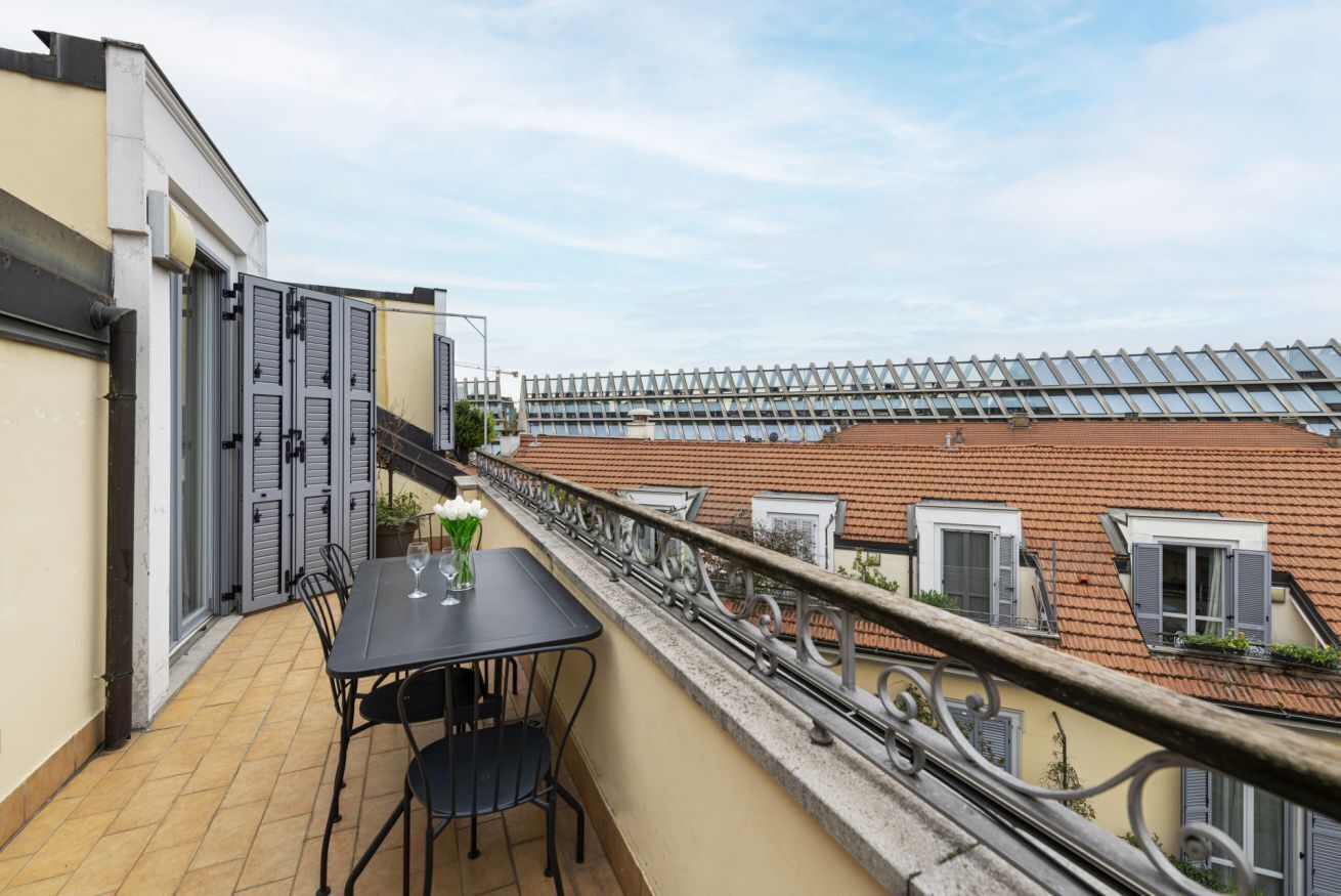 A balcony with a table and chairs, a potted plant, and a view of adjacent buildings with red-tiled roofs and windows with shutters.