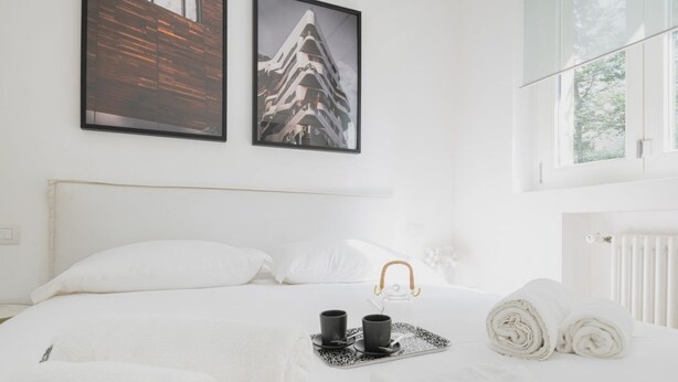A hotel room with a neatly made bed, two framed pictures on the wall, a window with a view of trees, and a tray with two cups and a kettle. - Easylife - Elegance and Comfort in the Isola area (Milano)