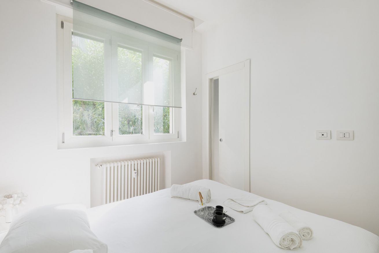 A minimalist hotel room with a large bed, a window with blinds, a radiator, and a door.