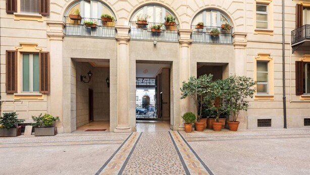 Fachada de un edificio con un sendero de baldosas que conduce a una puerta, flanqueada por macetas y un balcón con cestas de flores colgantes.
