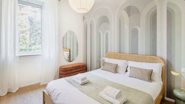 A bedroom with a large bed, a bedside table, a mirror, and a window with curtains.