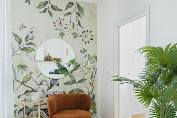 Una habitación con papel tapiz floral, un espejo redondo, un sillón marrón, una mesita auxiliar y una planta grande en maceta.