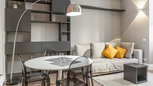 A modern living room with a round dining table, clear chairs, a sofa with cushions, a floor lamp, and a wall-mounted light fixture. - Easylife - Luxury and Comfort in Brera district (Milano)