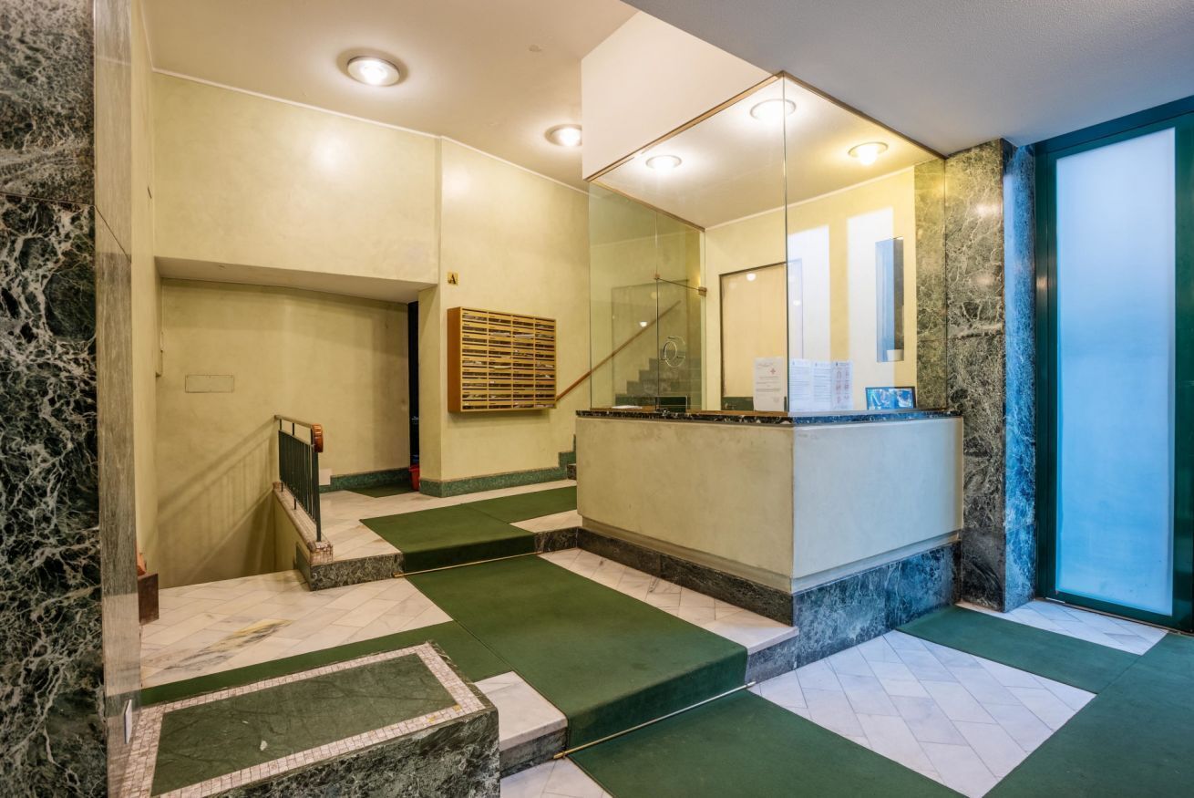 Eine Hotelhalle mit einem Marmor-Rezeptionsschalter, einer Treppe mit grünem Teppich und einer Glastür zu einem weiteren Bereich.