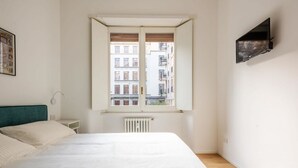 A bedroom with a large bed, a window with an open shutter, a radiator below, and a mounted TV on the wall. - Easylife-Spacious and comfortable apartment in the Duomo (Milano)