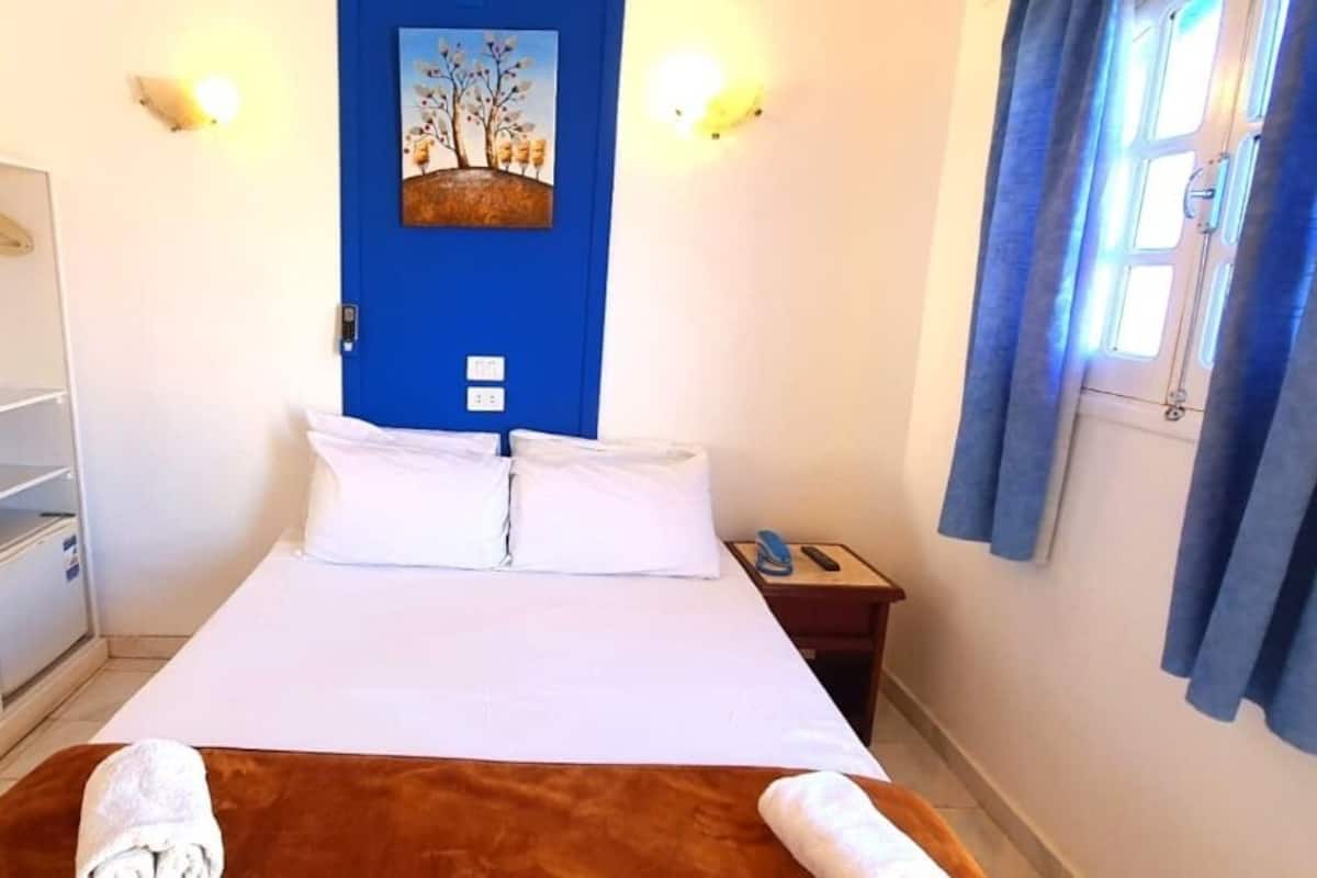 A hotel room with a bed, a wardrobe, a mirror, a painting on the wall, and a window with curtains.