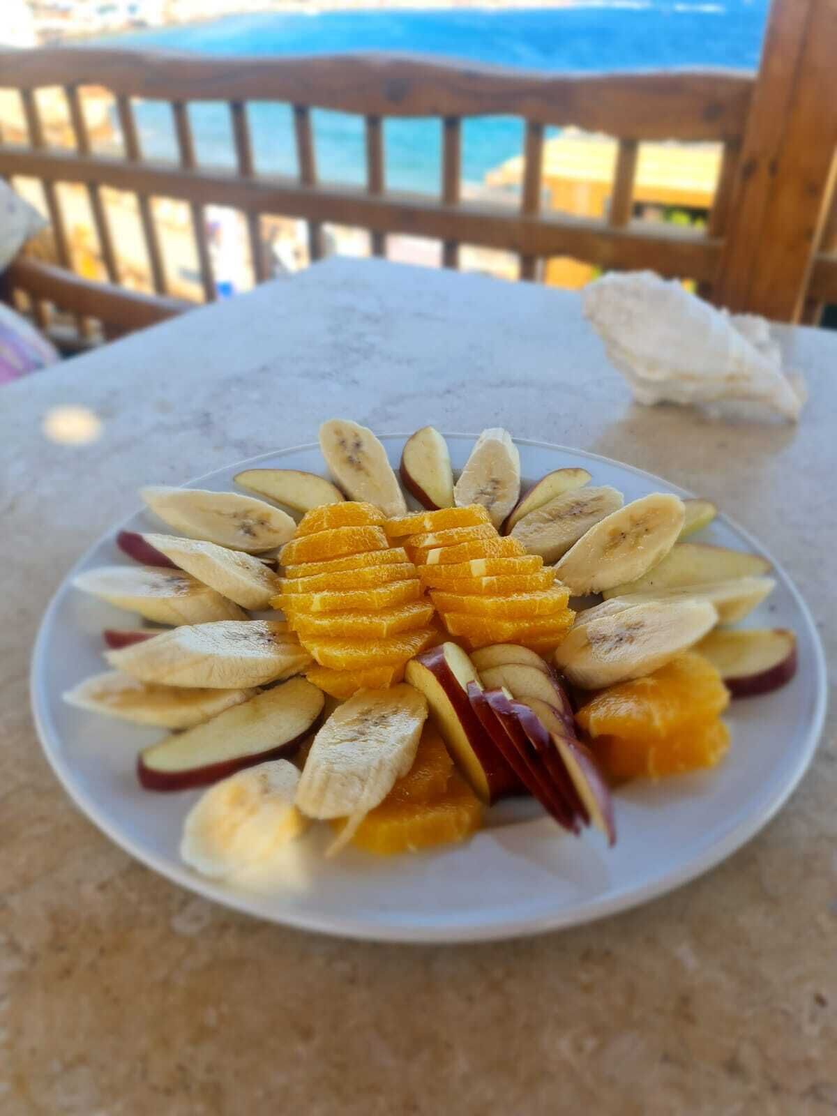 A plate of sliced fruits on a table with a wooden railing and a view of the sea in the background.