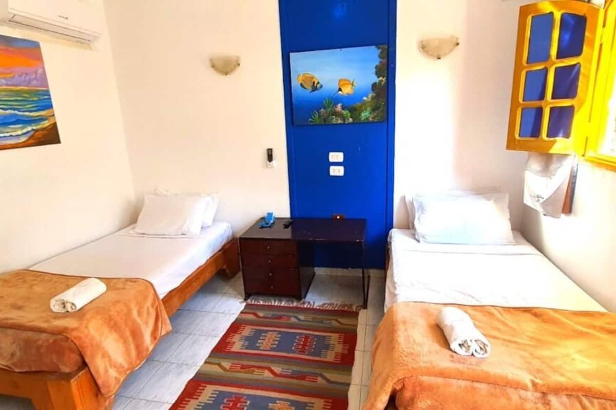 A small hotel room with two single beds, a small table, a blue door, and a painting of a sunset on the wall.