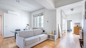 Ein geräumiges Wohnzimmer mit grauem Sofa, Holzboden, einem kleinen weißen Tisch mit Dekoration und einer Holzkommode mit Schubladen.