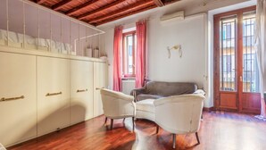 Une pièce avec un plafond en bois, une armoire blanche aux poignées dorées, un canapé gris et deux fauteuils blancs.