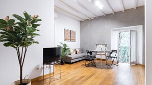 Una sala moderna con piso de madera, televisor negro, sofá gris, mesa de comedor con sillas y una planta grande en maceta.
