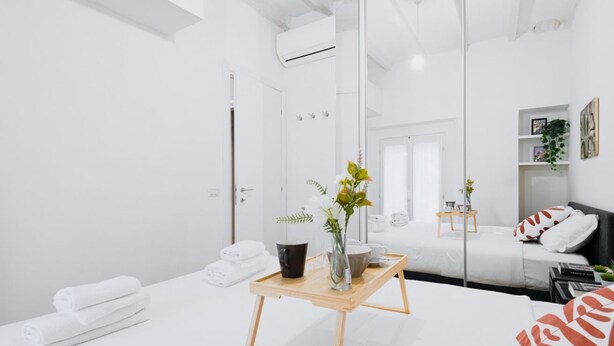 Una habitación de hotel moderna con un espejo grande, una mesita de noche de madera con un jarrón y una bandeja, una cama con ropa blanca, y una pequeña mesita de noche con una planta.
