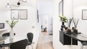 A modern, minimalist living room with a glass dining table, black chairs, and a black sofa. There are framed artworks on the walls and a plant on the table.