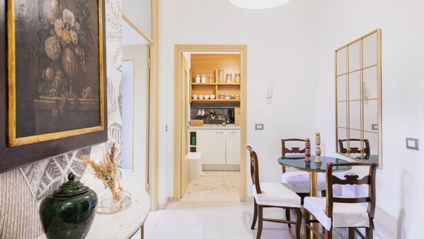 A dining area with a glass table and wooden chairs, a kitchen in the background, and a framed painting on the wall. - Easylife - Classy abode in the Conciliazione area (Milano)