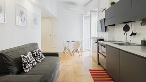 Een modern appartement met een grijze bank, een eethoek met een witte tafel en stoelen, een keuken met grijze kastjes en een rode vloerkleed.