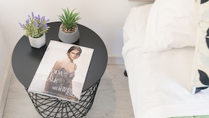 Une chambre à coucher avec un lit, une petite table d’appoint ronde et noire sur laquelle repose un magazine, et deux plantes en pot.