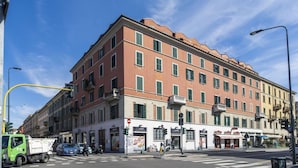 Un hôtel à plusieurs étages, avec une façade rouge et des volets verts, situé à un carrefour, près d’un passage piéton et de feux de signalisation.