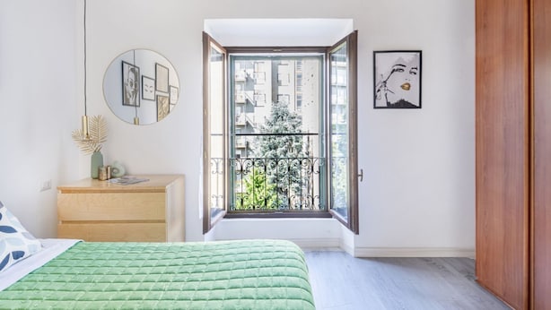Une chambre avec un lit vert, une commode en bois, un miroir et une fenêtre donnant sur un balcon.