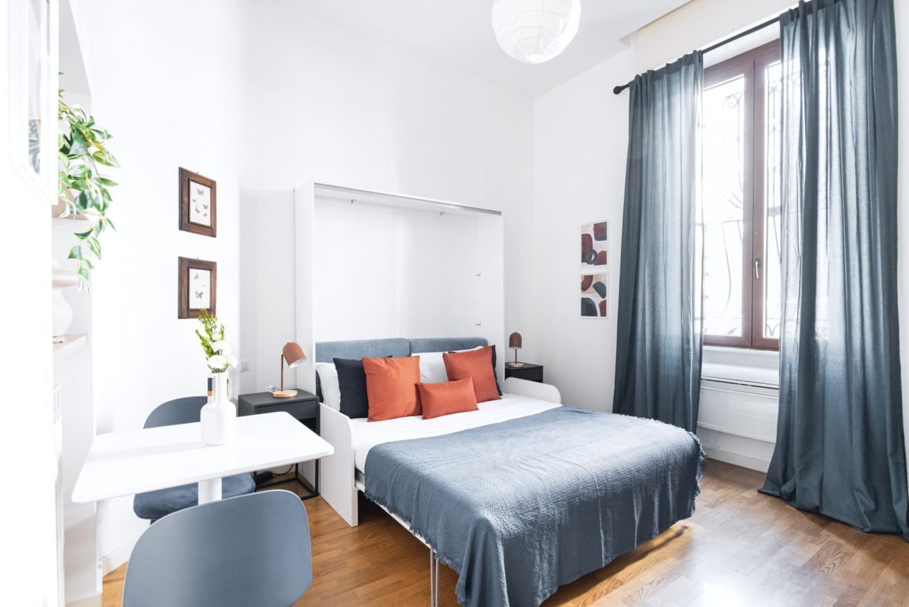 Une chambre à coucher moderne avec un lit, des tables de chevet, un bureau et une chaise. Il y a des rideaux à la fenêtre et une plante sur le bureau.