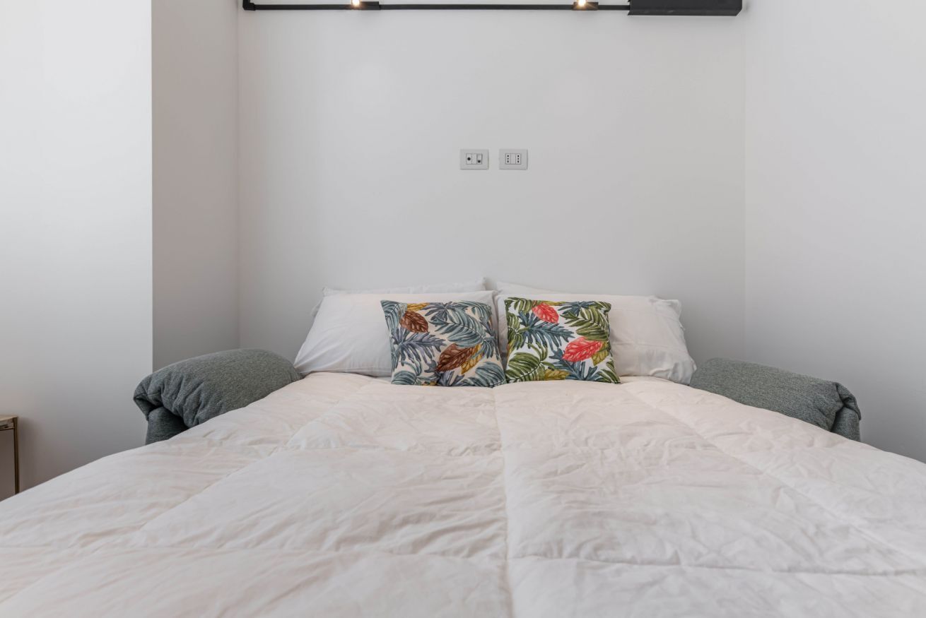 Un lit bien fait, avec une literie blanche et deux coussins décoratifs ornés d’un motif de feuilles tropicales.