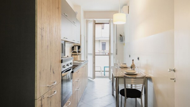 A narrow kitchen with wooden cabinets, a stove, and a dining area with a table and chairs. - Easylife - Charming flat 5 min from Centrale (Milano)