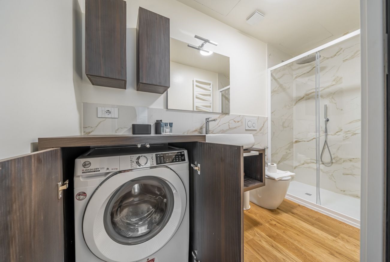 Une salle de bain compacte dotée d’un lave-linge intégré, d’une douche aux murs en marbre et d’un meuble-lavabo en bois.