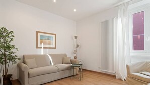 Una sala de estar con un sofá color beige, una mesita, una lámpara de pie, una planta en maceta y un ventanal con cortinas blancas.