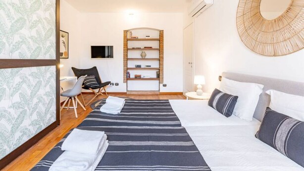 Una habitación de hotel con una cama, una silla, un estante con libros, un televisor montado en la pared y un espejo redondo.