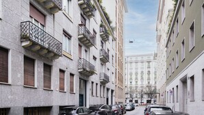 A narrow urban street lined with multi-story buildings, featuring balconies with plants, parked cars, and a clear sky. - Easylife - Gorgeous flat in the Design District (Milano)