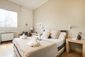 Une chambre d’hôtel moderne, dotée d’un grand lit, de tables de chevet, d’un bureau et d’une fenêtre donnant sur des arbres sans feuilles.