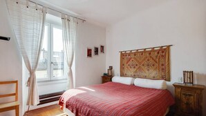 A bedroom with a bed, a wooden bench, a nightstand, a patterned tapestry, and a window with curtains. - Easylife - Attico Duplex Chinatown (Milano)