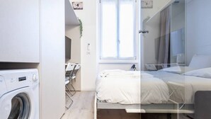 A compact hotel room with a washing machine, a bed, and a window providing natural light. - Easylife - Cozy studio apartment in Porta Venezia (Milano)