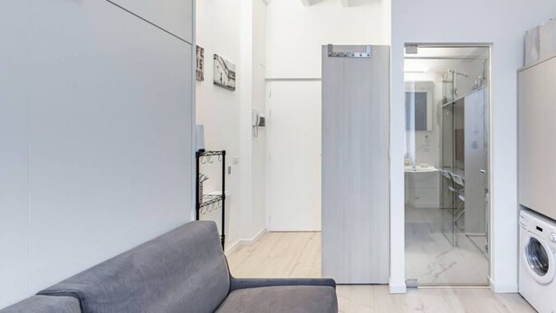 A modern living room with a grey sofa, a washing machine, and a glass shower enclosure. - Easylife - Cozy studio apartment in Porta Venezia (Milano)