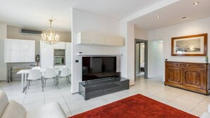 A modern living room with a fireplace, a dining area with white chairs, and a kitchen with white cabinets.