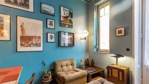 Een kamer met een blauwe muur, een groot raam, een bank, een tafel en verschillende lijstwerken.