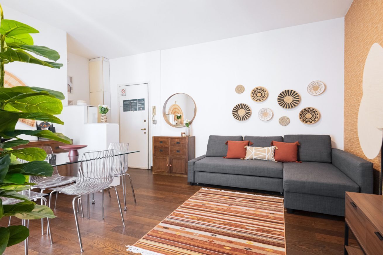 Ein modernes Wohnzimmer mit einem grauen Sofa, einem Holztisch und einem Streifen Teppich. Es gibt einen Essbereich mit einem Glastisch und Metallstühlen. An den Wänden sind außerdem Wandbilder und ein Spiegel zu sehen.