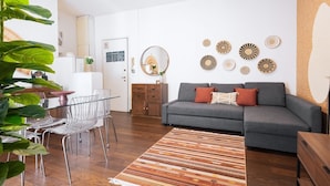 Una sala de estar moderna con un sofá gris, una mesa de centro de madera y un tapete a rayas. Se observa un comedor con una mesa de vidrio y sillas de metal. También hay arte decorativo en la pared y un espejo.