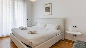 Un lit bien fait, avec du linge de lit blanc et deux serviettes enroulées, une table de chevet avec une lampe, un tableau encadré au mur et une fenêtre avec des rideaux légers.